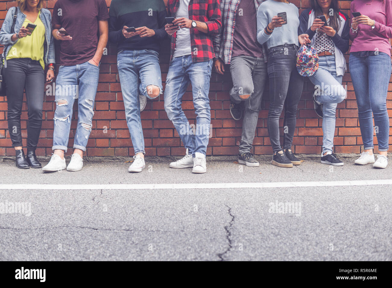 Group of eight teen friends millenials boys and girls watching smart mobile phones in front of brick wall background. Concept of youth, tech, social a Stock Photo