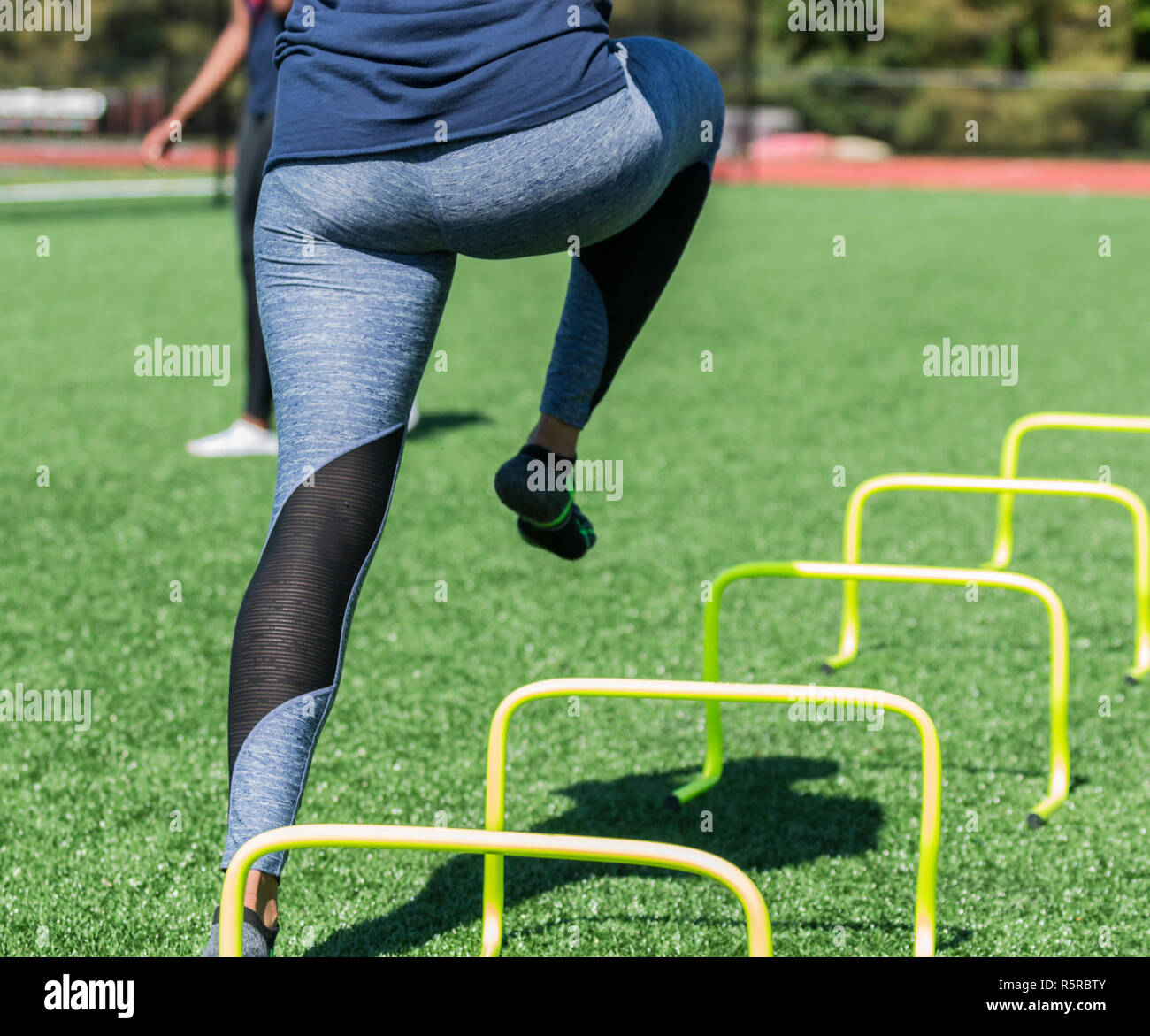 A female high school athlete performs running drills over yellow mini banana hurdles on a turf field with no shoes on, only socks in blue spandex. Stock Photo