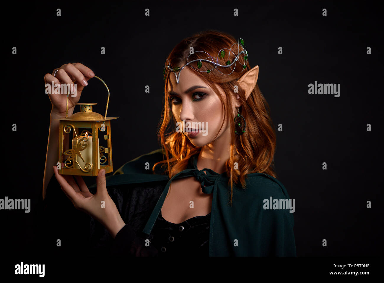 Portrait of charming mythical creature with red hair, in black dress and dark green cloak. Young beautiful elf with tiara on head holding in her hand golden lantern with white candle inside. Stock Photo