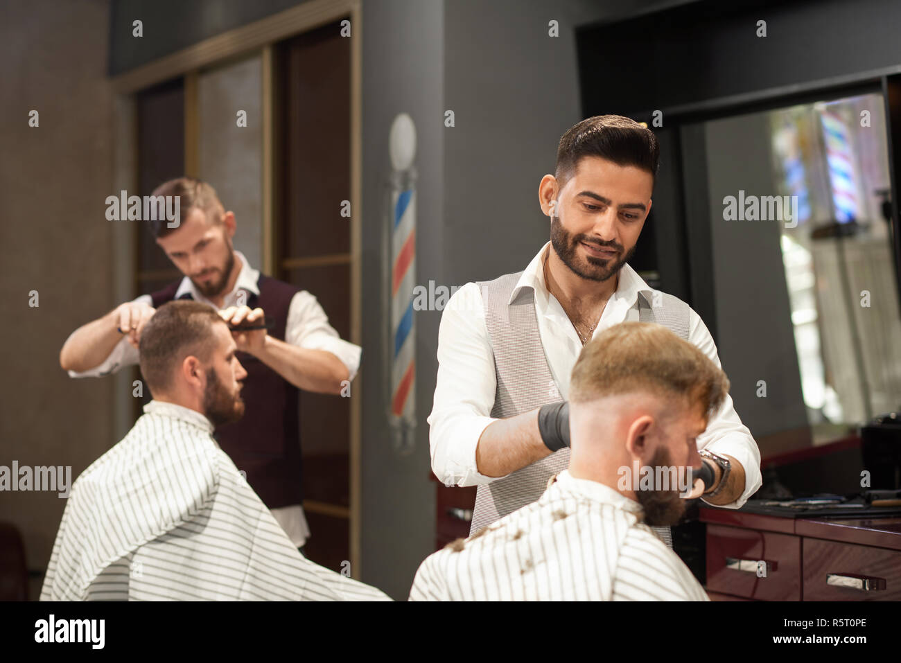 View from side of professional barber servicing male client sitting in chair in barbershop. Stylish hairdresser in uniform and protective gloves cutting hair. Concept of hairstyling and shaving. Stock Photo