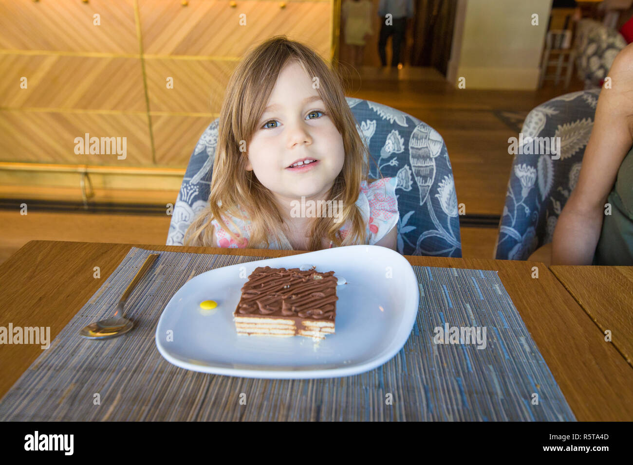 three years old child looking with a portion of chocolate cake in white dish over placemat sitting next to woman in restaurant Stock Photo