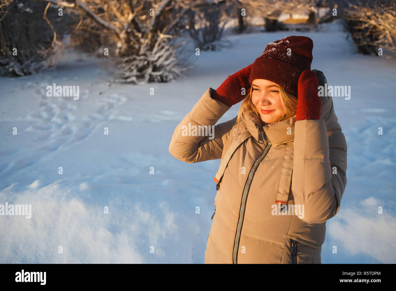 Pretty young caucasian woman in red mittens and red winter hat enjoying good sunny winter day in snowy nature. She holding her hat and looking to the sun. Good mood, weather, and winter love concept. Stock Photo