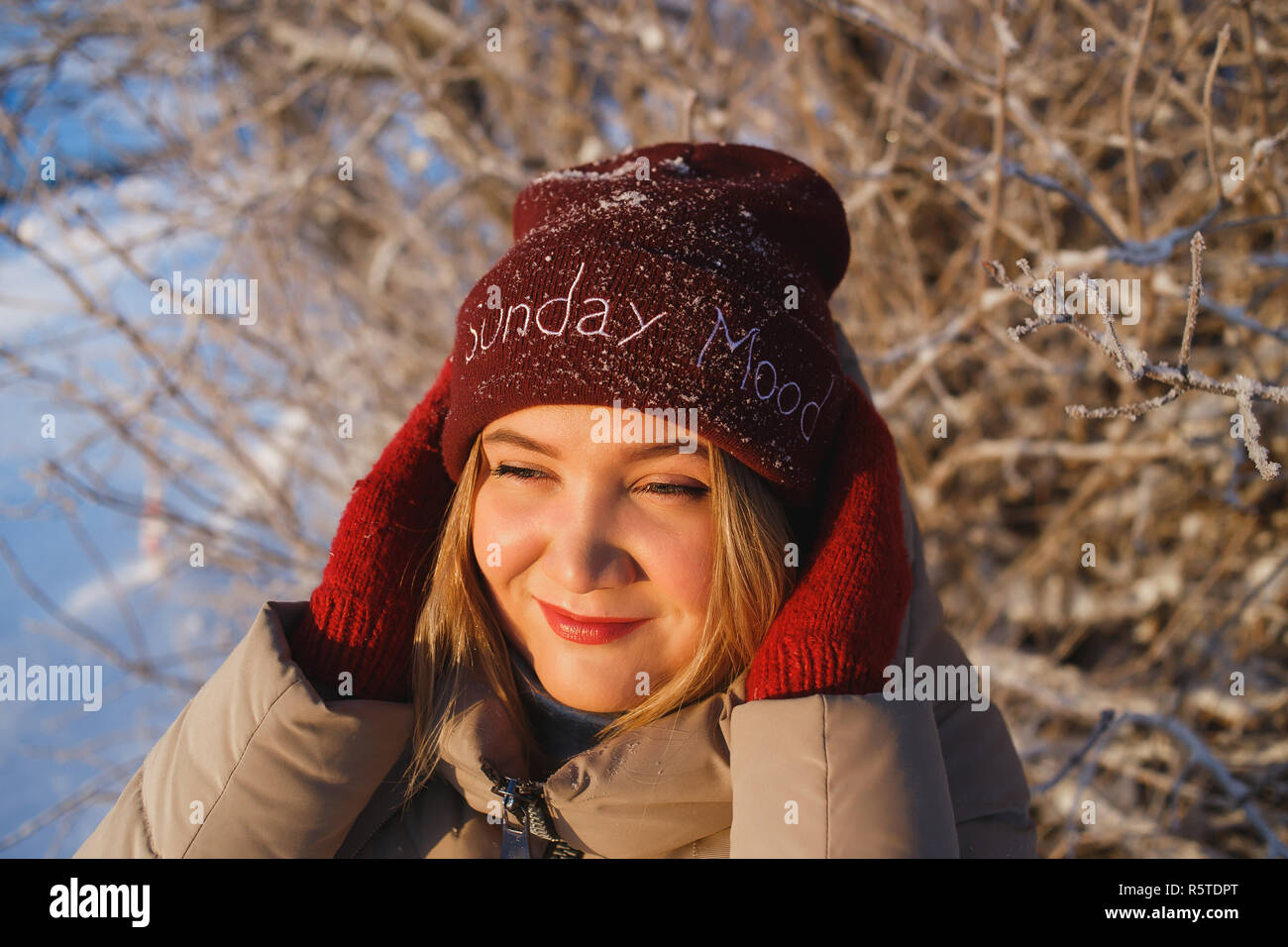 Pretty young caucasian woman in red mittens and red winter hat enjoying good sunny winter day in snowy nature. She holding her hat and looking to the sun. Good mood, weather, and winter love concept. Stock Photo