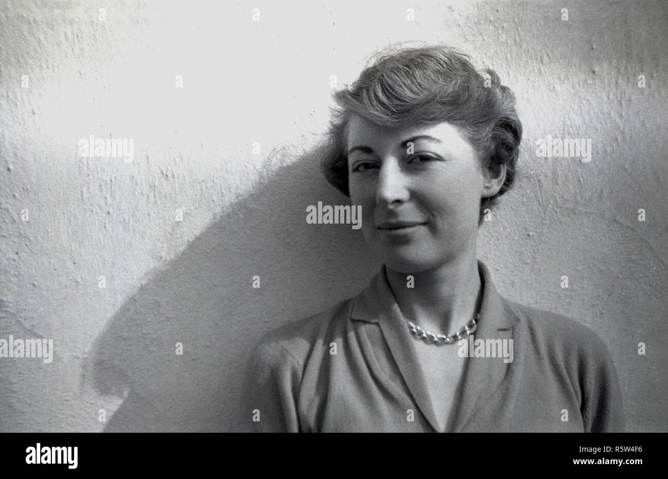 1950s, historical, attractive youg English lady with short wavy hair and wearing a necklace and woollen top standing against a wall, England, UK. Stock Photo