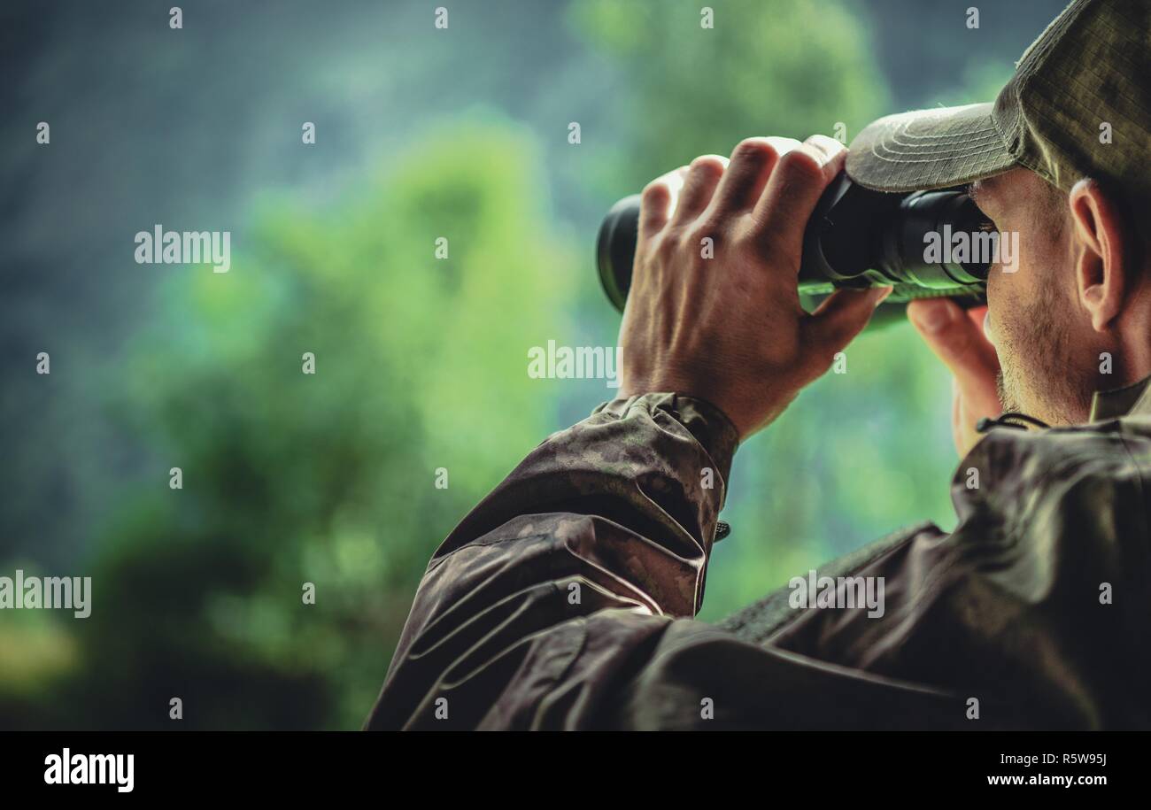 Caucasian Hunter in Camouflage with Binoculars Spotting Wildlife in the Remote Place. Hunting Theme. Stock Photo