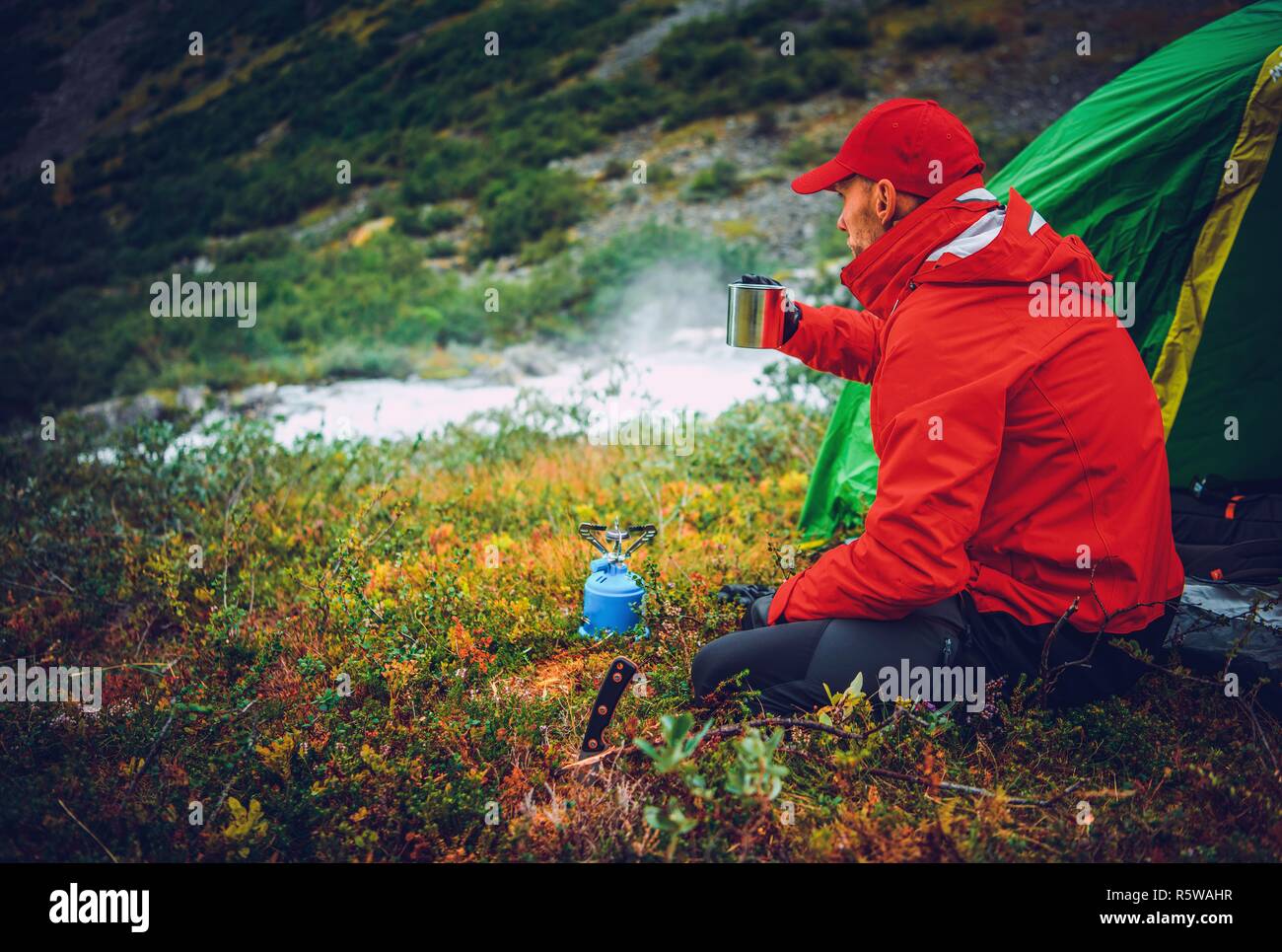 Caucasian Men Warming Up by Drinking Hot Tea in Front of His Tent. Wilderness Campsite. Stock Photo