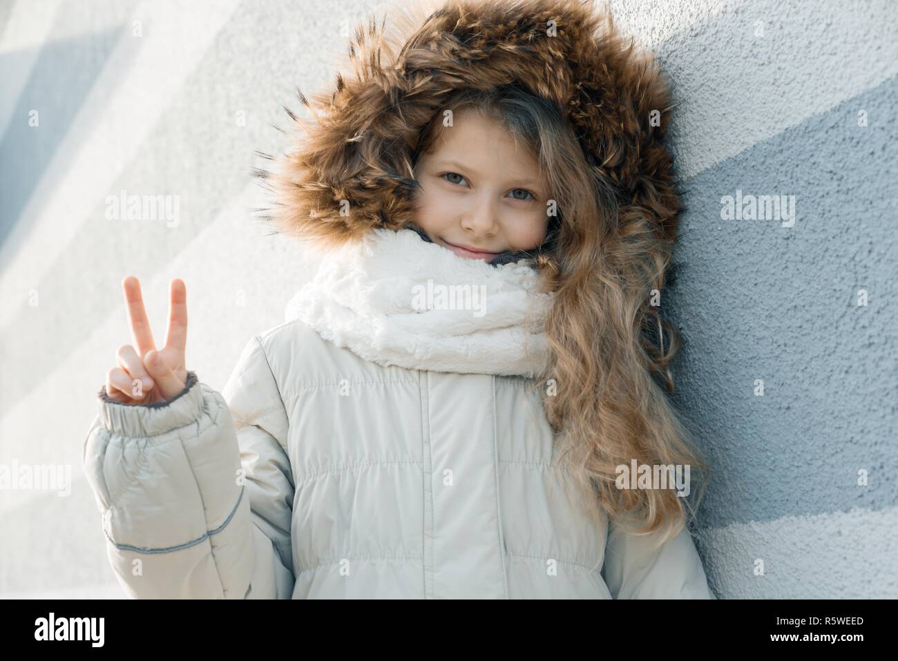 Close-up outdoor winter portrait of child, blonde girl with curly hair of 7, 8 years in fur hood, girl smiling showing victory sign. Stock Photo