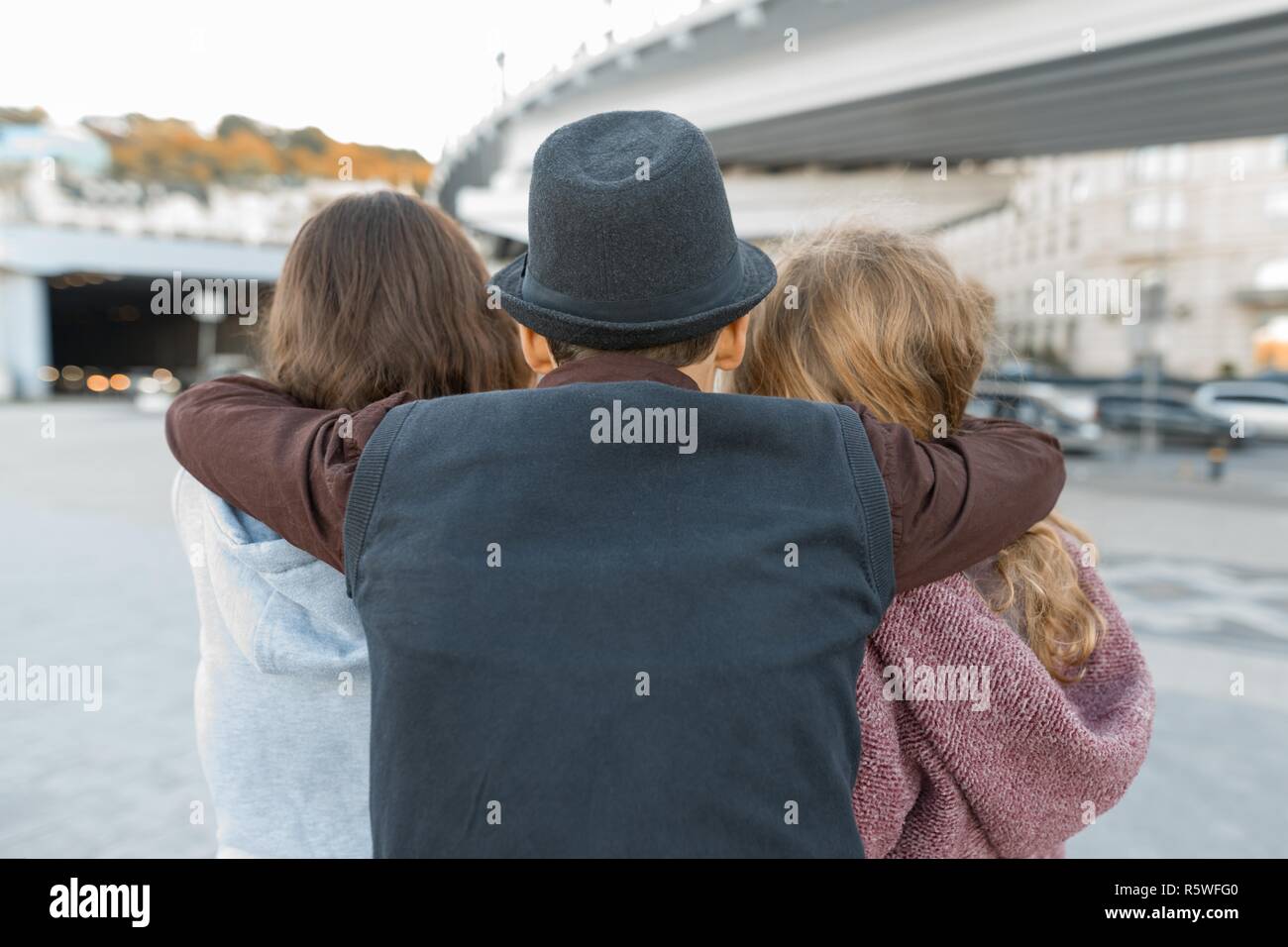 Three friends, view from the back of young teenager boy hugging two girls by the shoulders. Children look ahead, city life Stock Photo