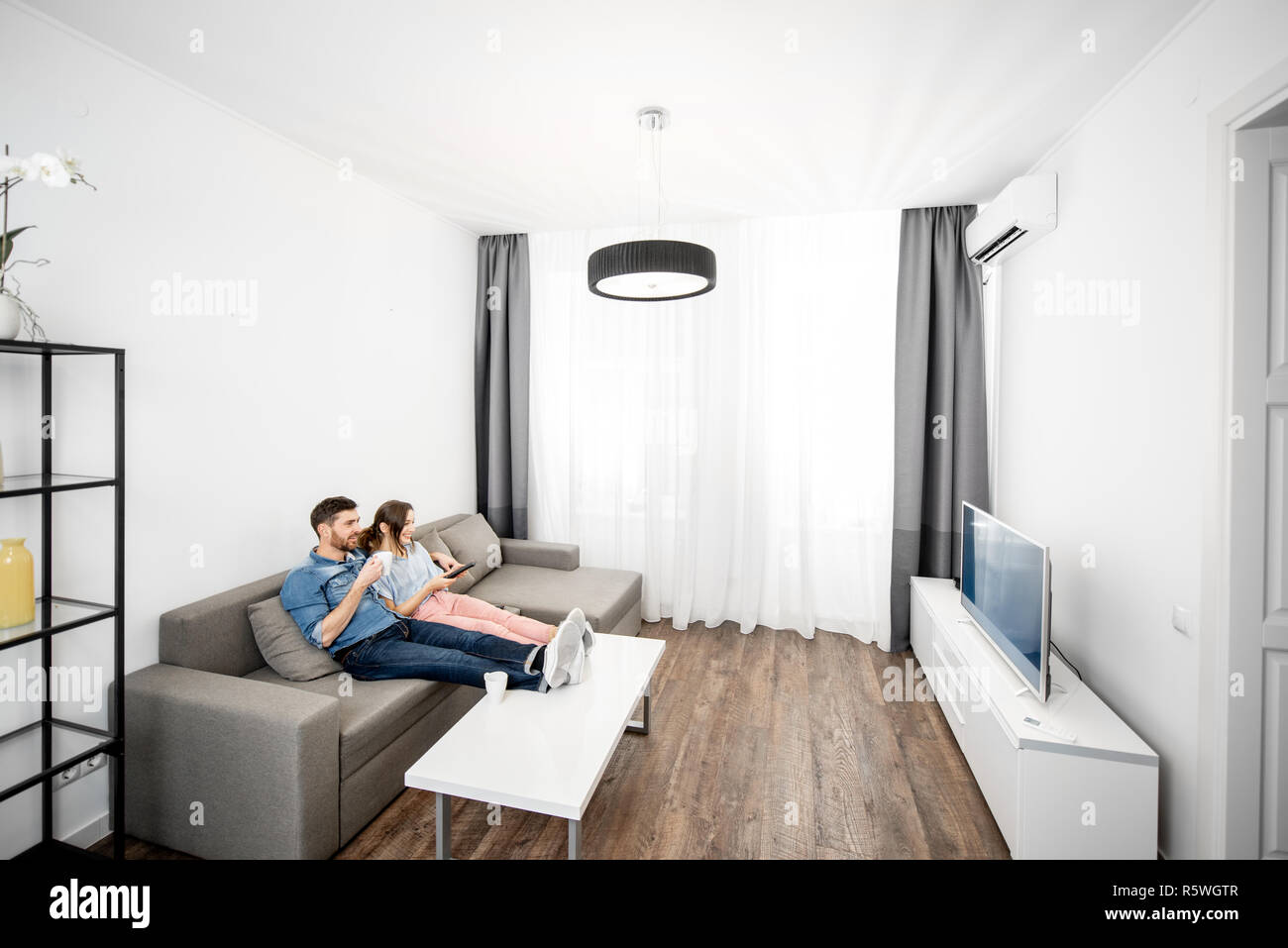 Young couple sitting together on the couch and watching TV at home. Wide interior view Stock Photo