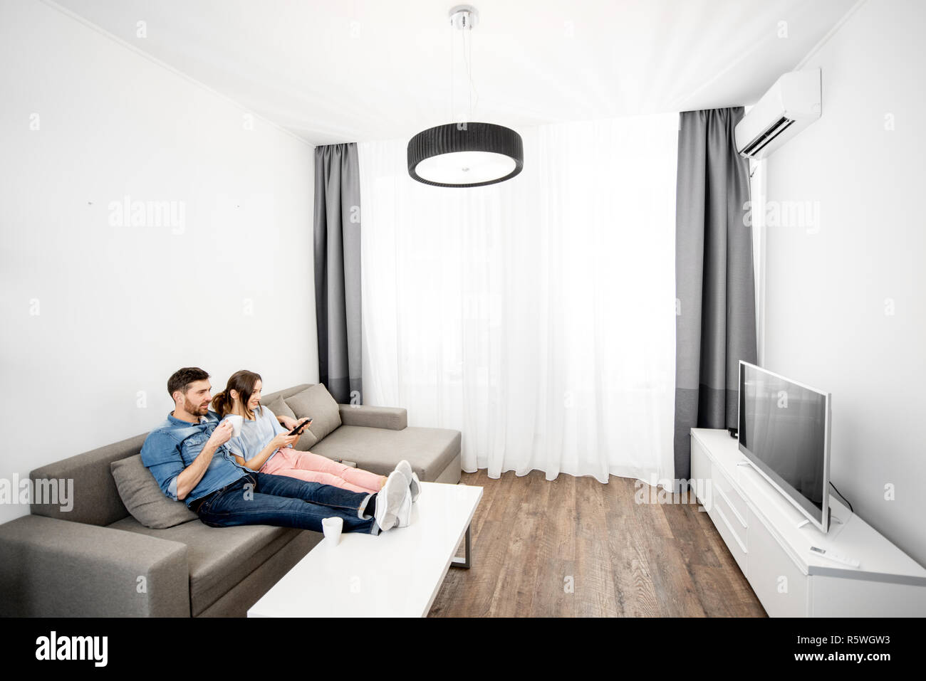 Young couple sitting together on the couch and watching TV at home. Wide interior view Stock Photo
