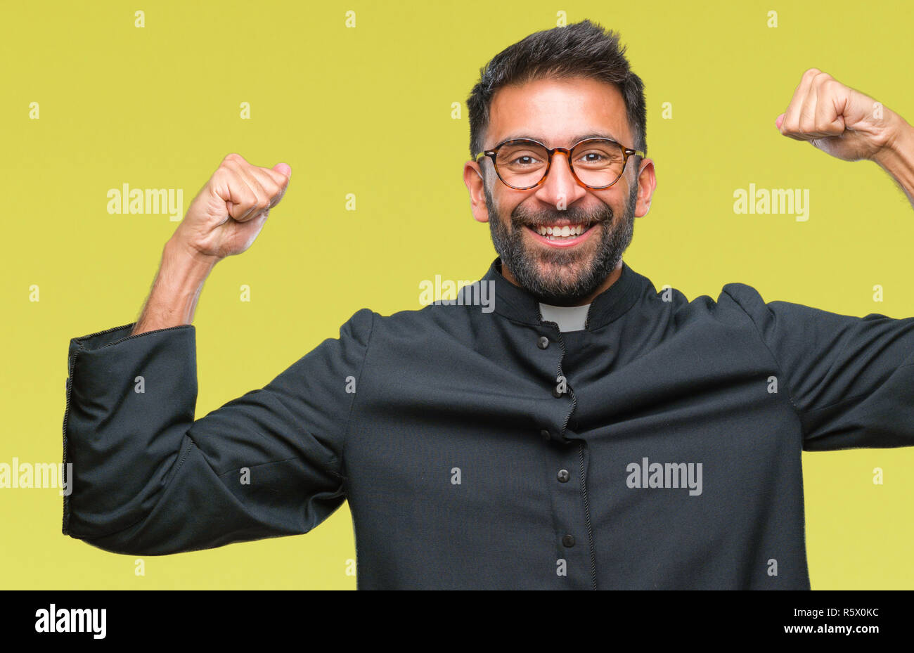 Adult hispanic catholic priest man over isolated background showing arms muscles smiling proud. Fitness concept. Stock Photo