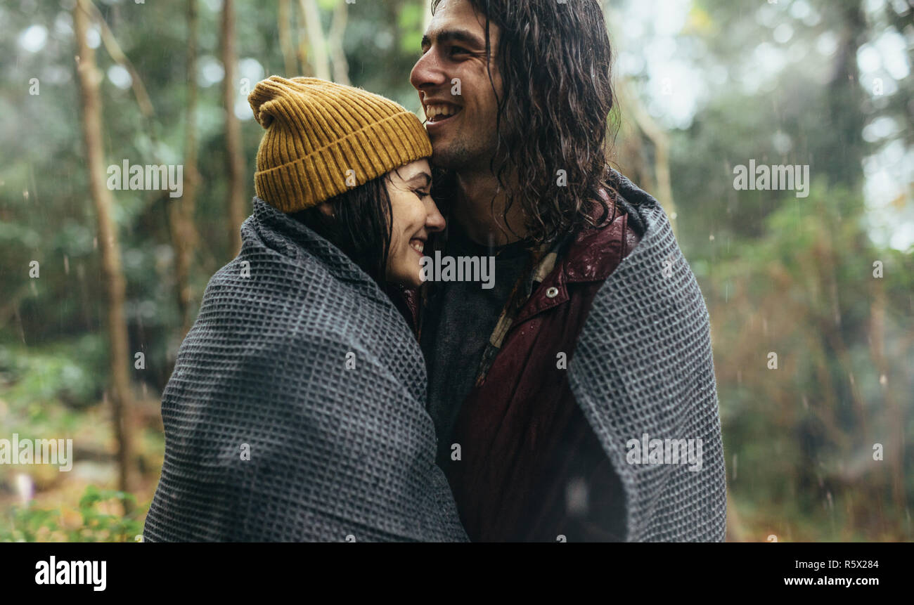 Couple in love standing under the rain wrapped in a blanket. Smiling man and woman embracing each other at the forest on a rainy day. Stock Photo