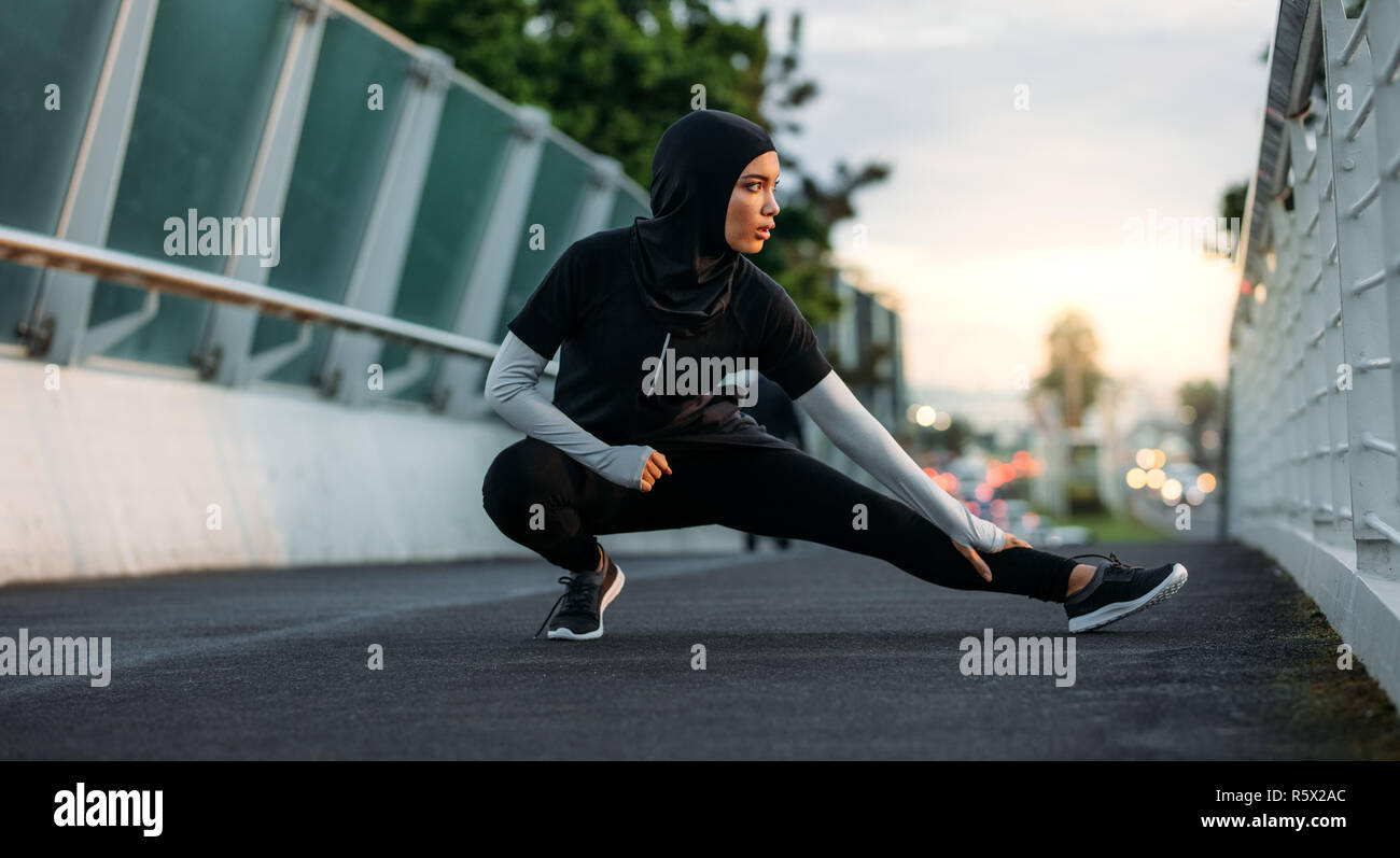 Hijab girl exercising on walkway bridge in early morning. Muslim woman wearing sports clothes doing stretching workout outdoors. Stock Photo