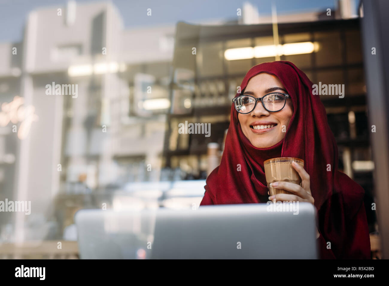 Beautiful muslim woman wearing hijab and eyeglasses relaxing at cafe having a cup of coffee and looking away with lovely smile on her face. Happy hija Stock Photo