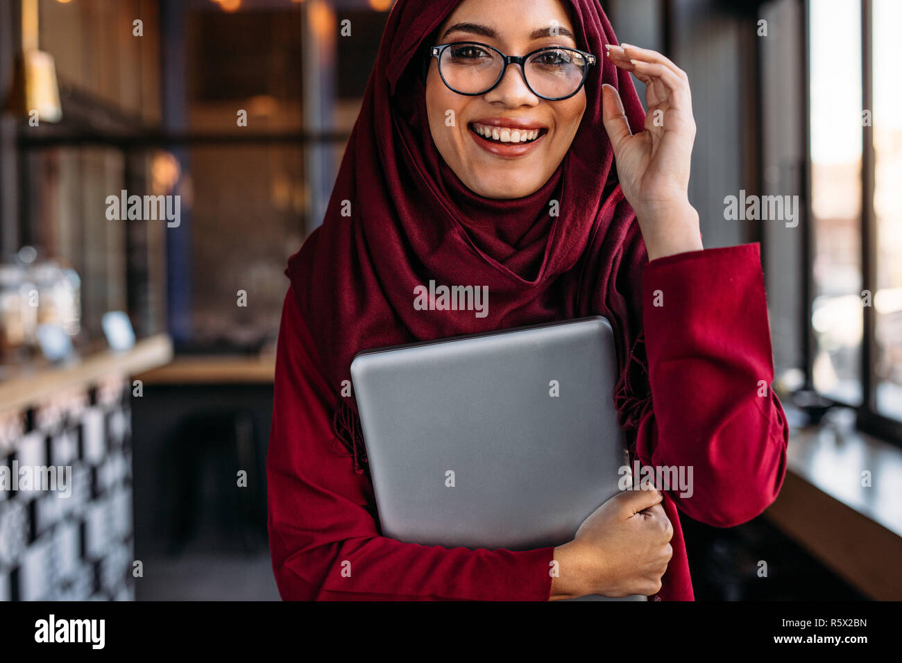 Beautiful muslim woman with laptop in hand adjusting her eyeglasses at coffee shop. Female in hijab at coffee shop. Stock Photo