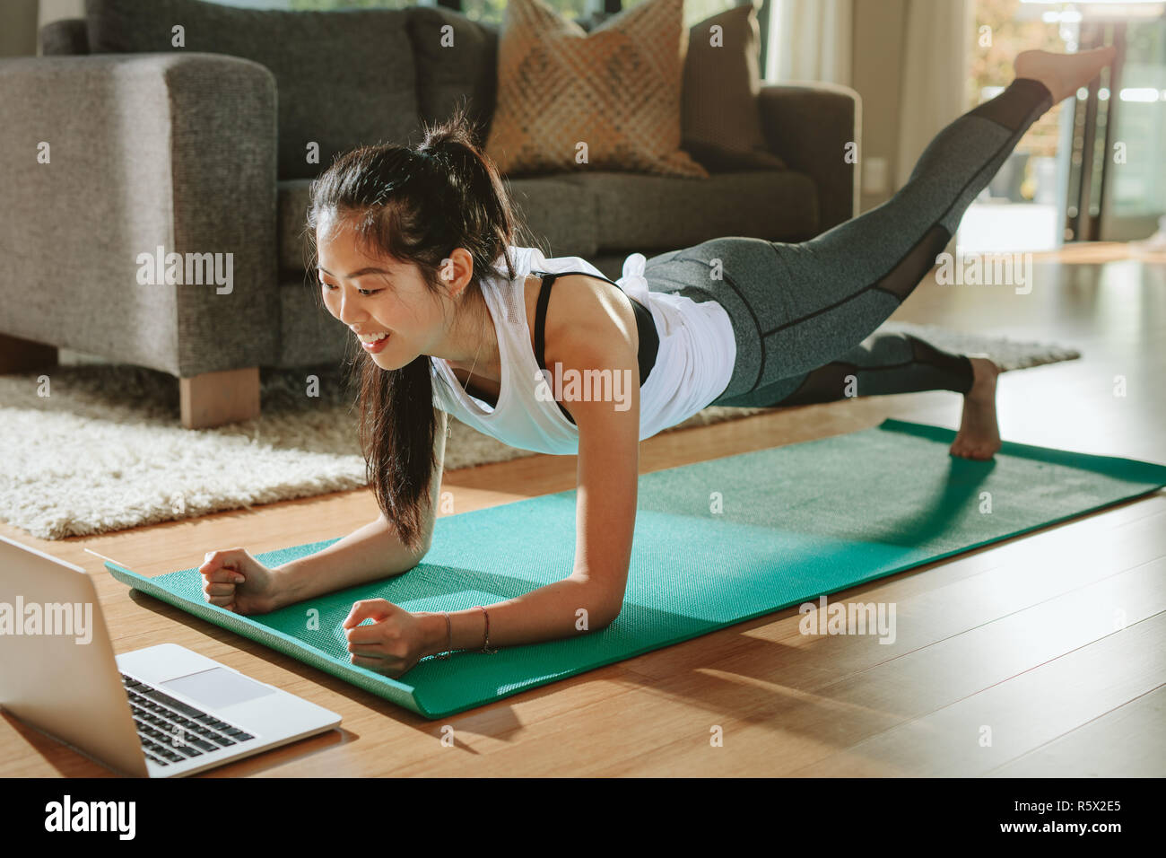 Smiling woman exercising at home and watching training videos on laptop. Chinese female doing planks with a leg outstretched and looking at laptop. Stock Photo