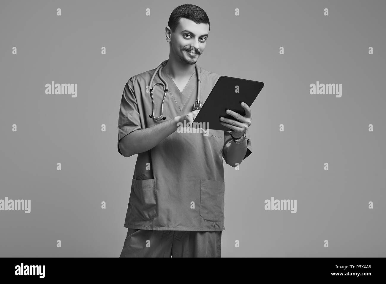 Portrait of handsome young confident arabic doctor with fancy mustache in blue holds tablet on gray studio background Stock Photo