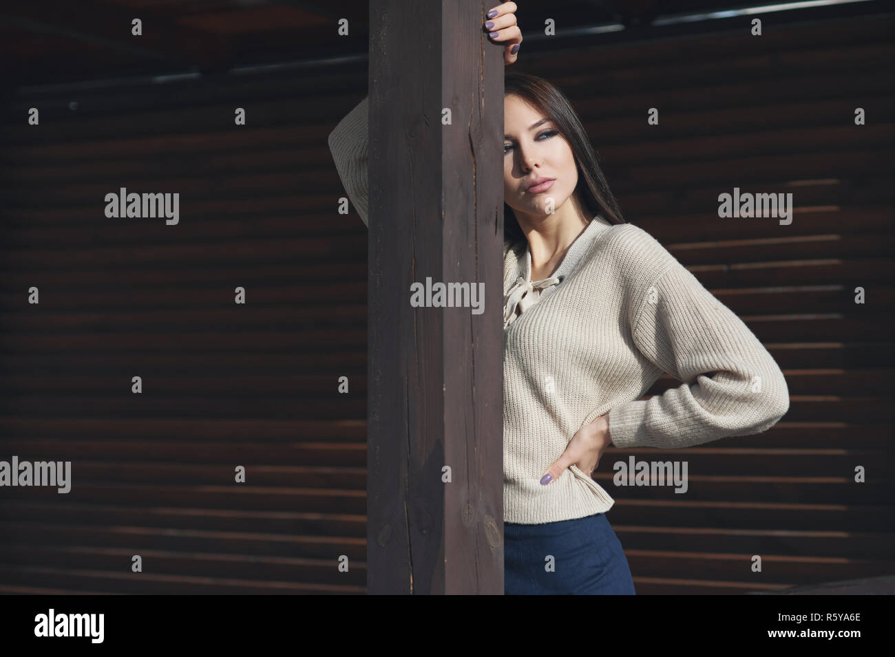 Portrait of a charming young woman in a knitted woolen beige sweater, on a wooden stall background. Hides half of face, and poses on camera Stock Photo