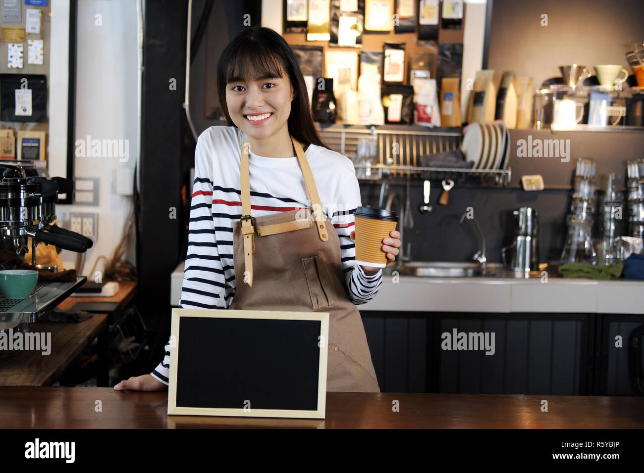 Smiling asian barista holding cup of coffee at counter in coffee shop. Cafe restaurant service, food and drink industry concept. Stock Photo