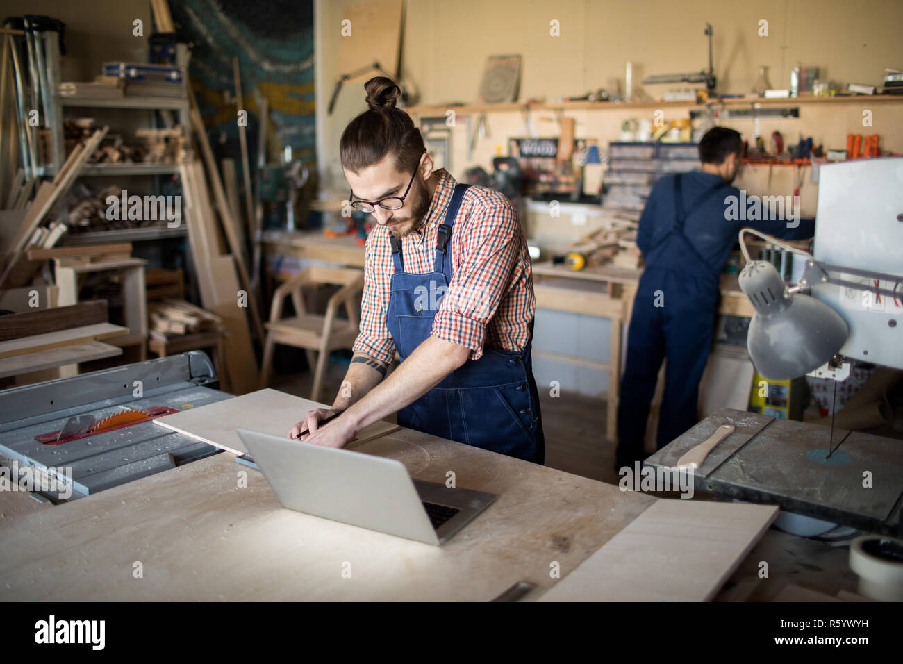 Portrait of two workers in carpenters workshop, focus on modern young man using laptop in foreground, copy space Stock Photo