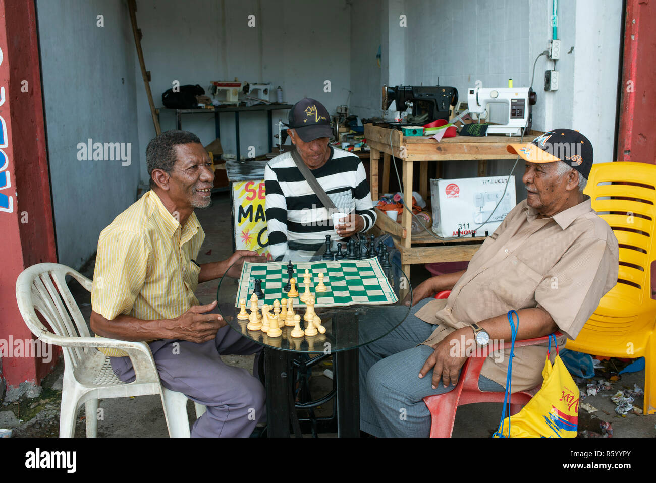 Senior local men playing chess outside a sewing machine repair shop. Daily life in Cartagena de Indias, Colombia. Oct 2018 Stock Photo