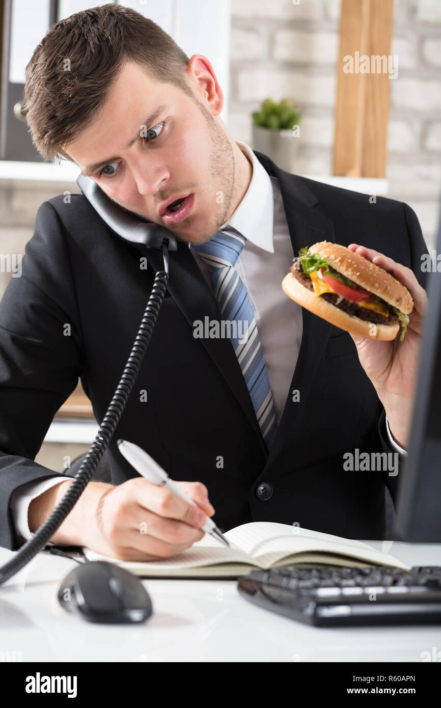 Business Man At Desk Eating Burger And Working Stock Photo