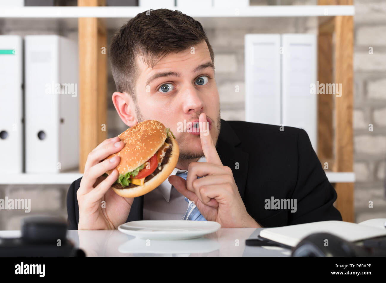 Hungry Young Businessman Holding Burger With Finger On Lips Stock Photo