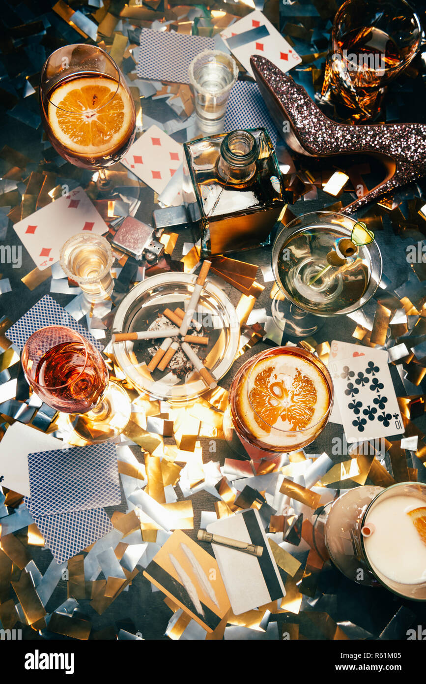 elevated view of alcoholic cocktails, playing cards, cigarettes, rolled banknote, credit cards and cocaine on table covered by golden confetti Stock Photo