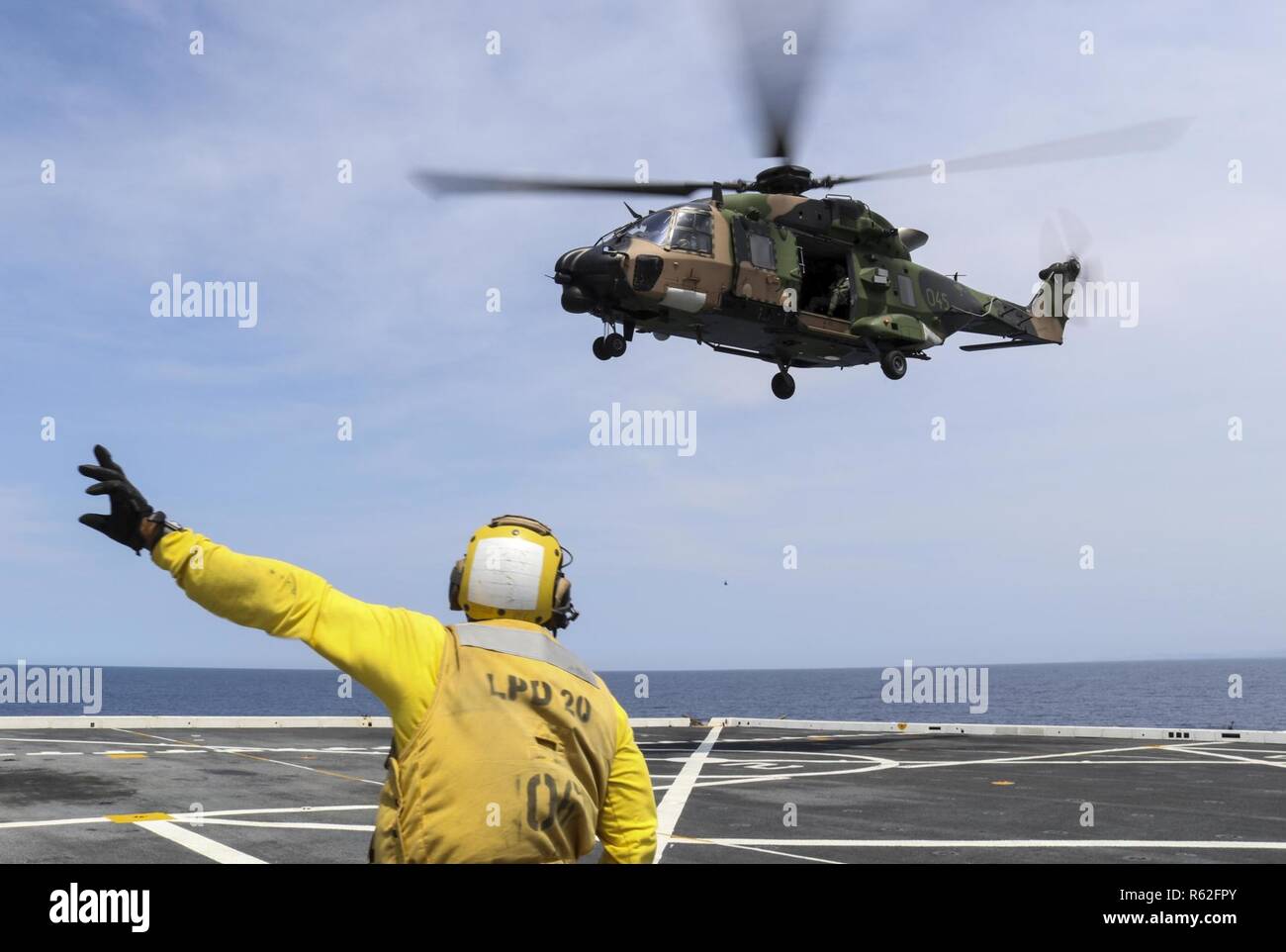 CORAL SEA (Nov. 19, 2018) Aviation Boatswain’s Mate (Handling) 3rd Class Dion Sanders, from New York, signals a MRH-90 Taipan helicopter, assigned to the Royal Australian Navy helicopter landing dock ship HMAS Adelaide (L01), as it takes off from the flight deck of the amphibious transport dock ship USS Green Bay (LPD 20) during cross deck flight operations. Green Bay, part of Commander Amphibious Squadron 11, is operating in the region to enhance interoperability with partners and serve as a ready-response force for any type of contingency. Stock Photo