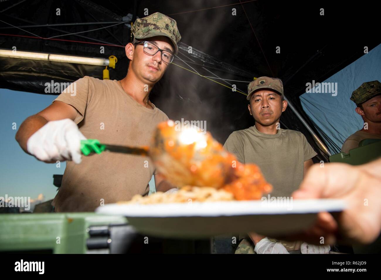 TINIAN, Commonwealth of the Northern Mariana Islands (Nov. 18, 2018) Construction Mechanic Construction Apprentice James Joyce, assigned to Naval Mobile Construction Battalion (NMCB) 1, Detachment Guam, from Pittsburgh, and Army Spc. Angelito Angala, assigned to the 303rd Maneuver Enhancement Brigade, from Honolulu, serve dinner to Sailors assigned to NMCB 1 during recovery efforts. Service members from Joint Region Marianas and U.S. Indo-Pacific Command are providing Department of Defense support to the Commonwealth of the Northern Mariana Islands' civil and local officials as part of the Fed Stock Photo