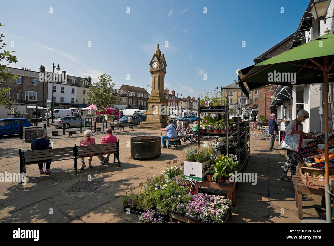 Plant stalls and people relaxing in the sunshine outside shops stores in summer Market Square Thirsk North Yorkshire England UK United Kingdom Britain Stock Photo