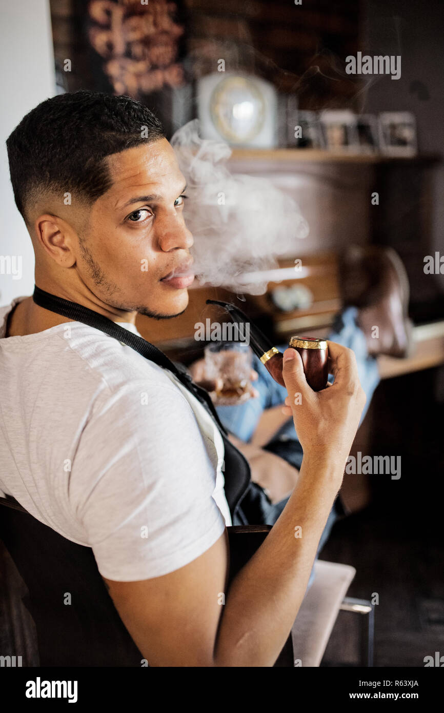 Young hispanic haidresser and hairstylist sitting in barber shop, smoking a pipe. Stock Photo