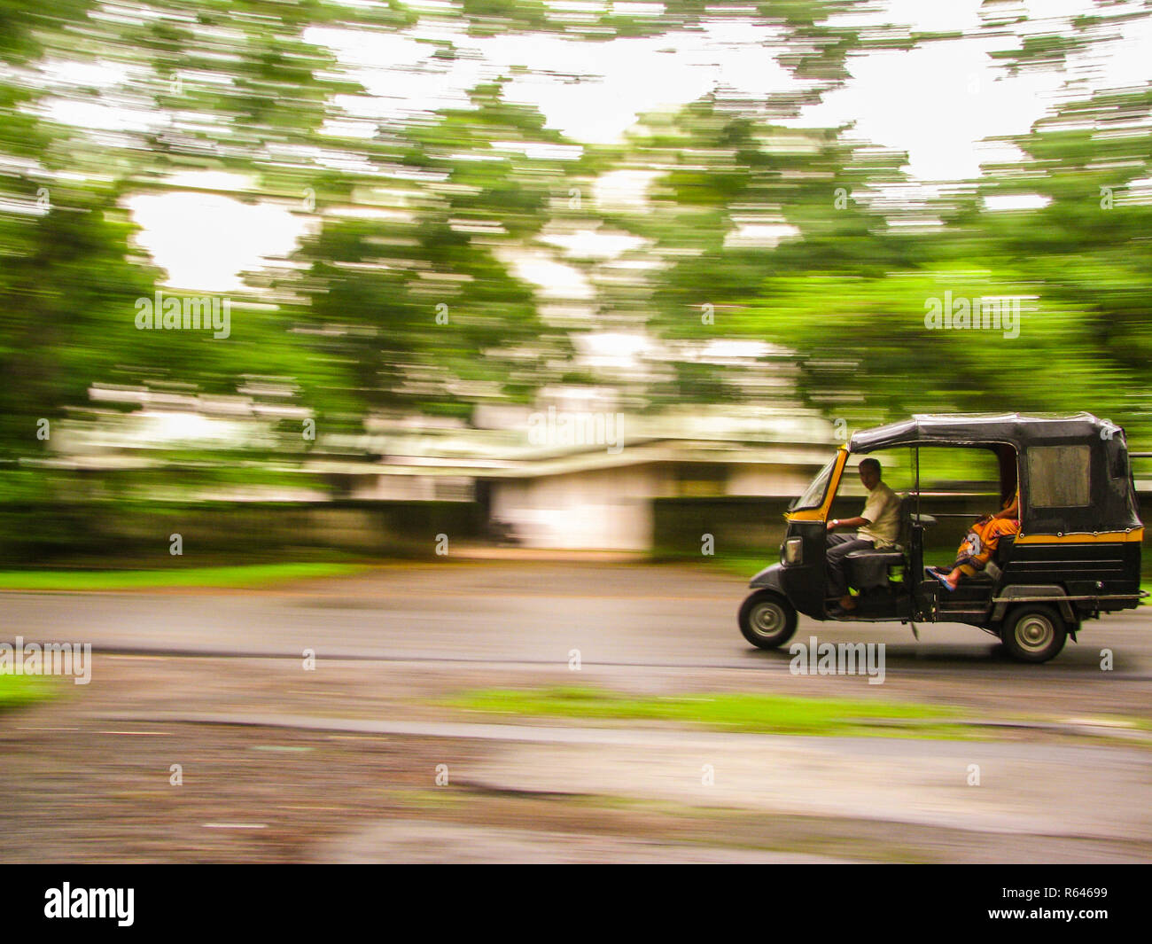 The photograph shows the panned shot of an Auto rickshaw speeding against trees in the background Stock Photo
