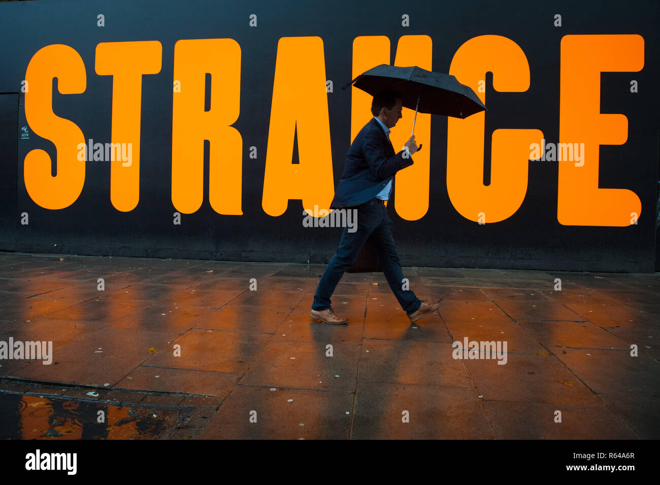 A man with an umbrella walks past an advertisement during a shower of rain in Aldwych, central