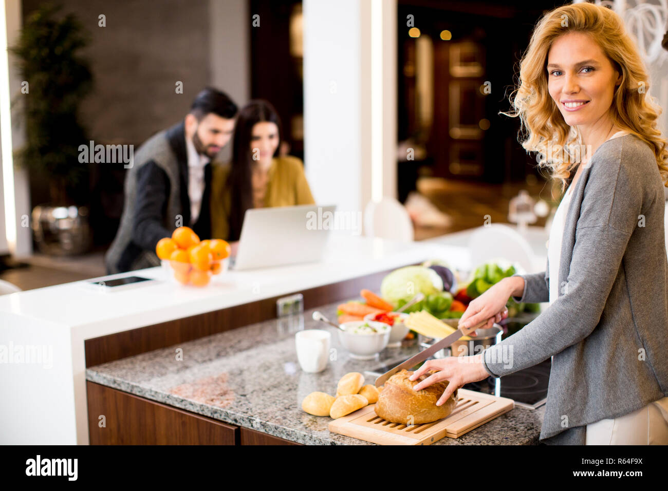Cheerful young woman preparing food while couple in background using laptop Stock Photo