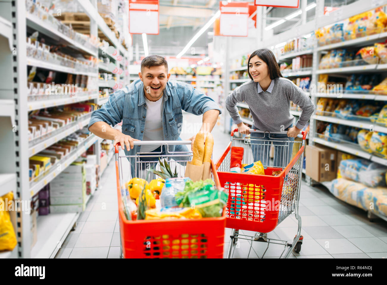 Playful couple ride on carts in supermarket, family shopping. Customers