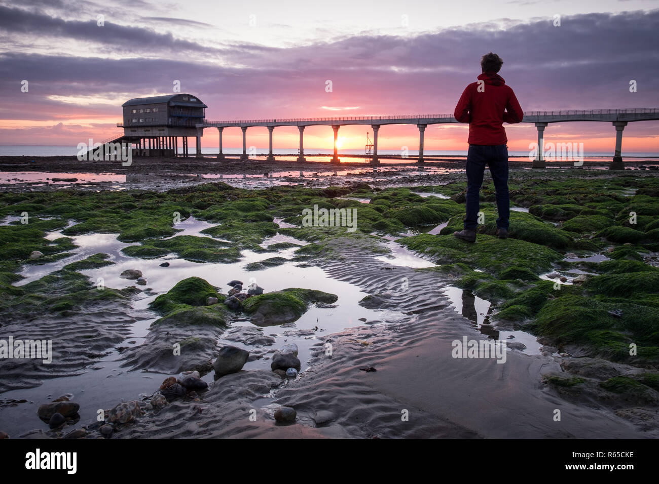 A man watches the sunrise over Bembridge RNLI Lifeboat station at Lane End Beach on the Isle of Wight Stock Photo