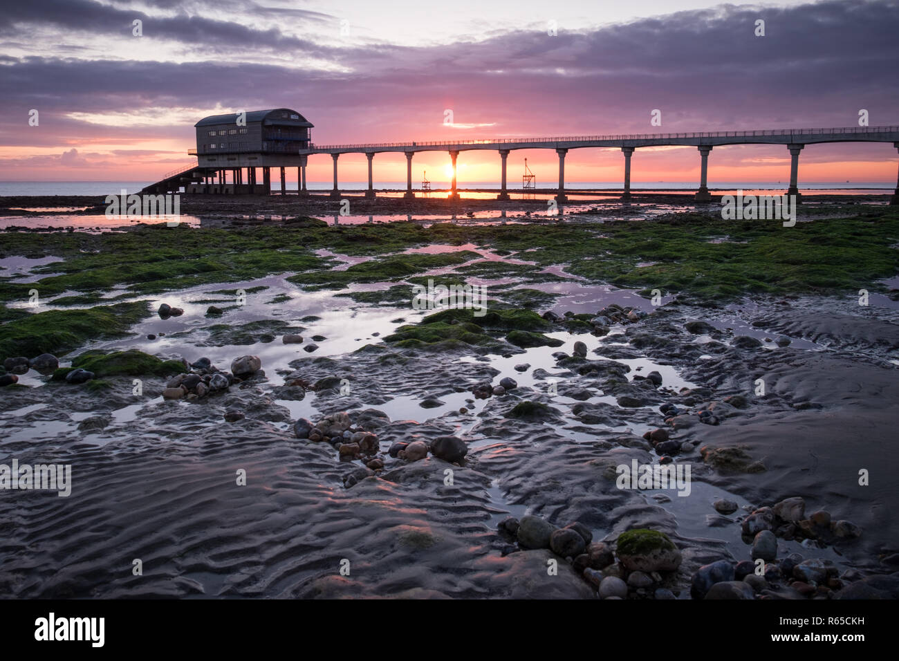 A man watches the sunrise over Bembridge RNLI Lifeboat station at Lane End Beach on the Isle of Wight Stock Photo