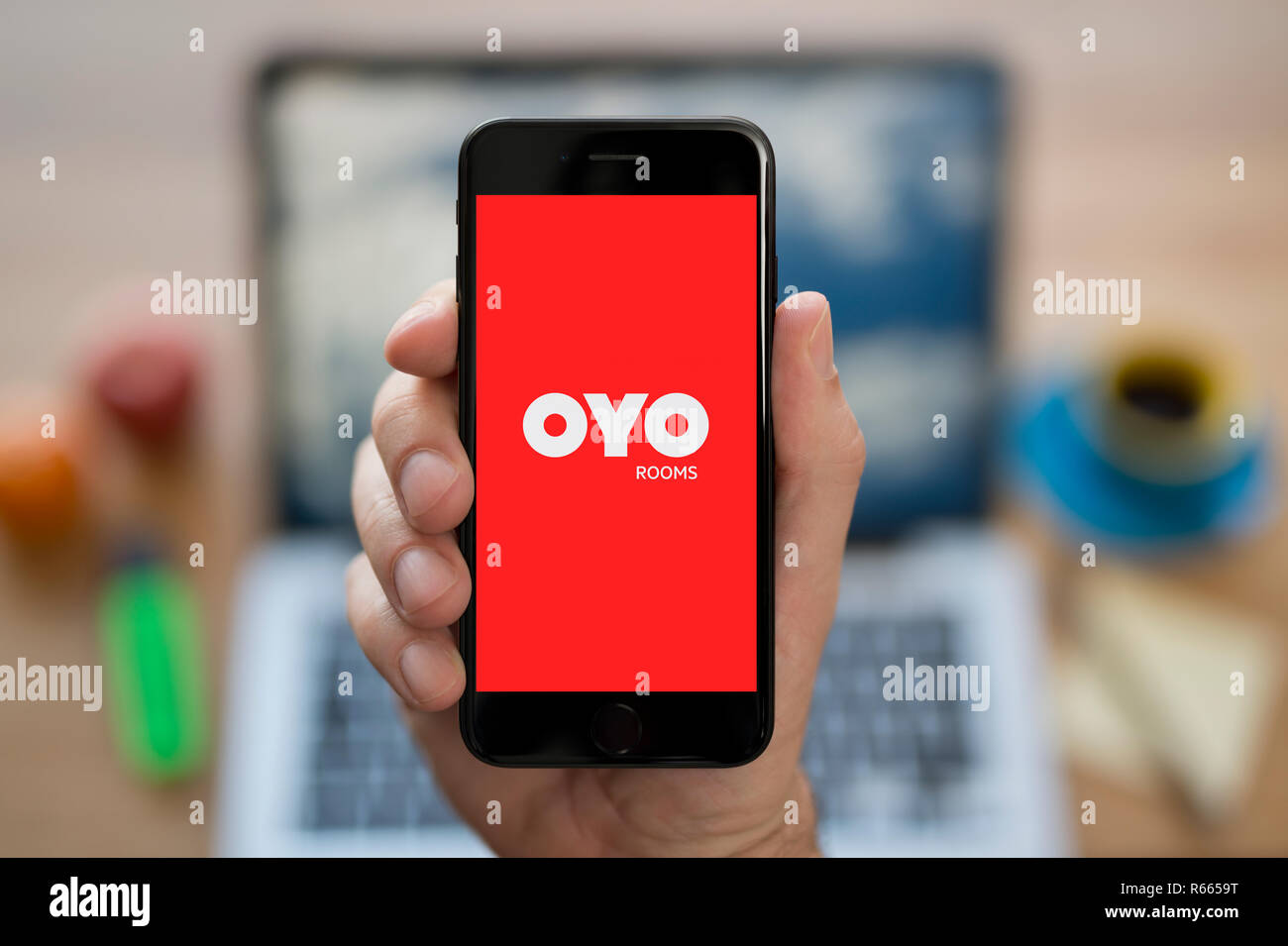 A man looks at his iPhone which displays the Oyo Rooms logo, while sat at his computer desk (Editorial use only). Stock Photo