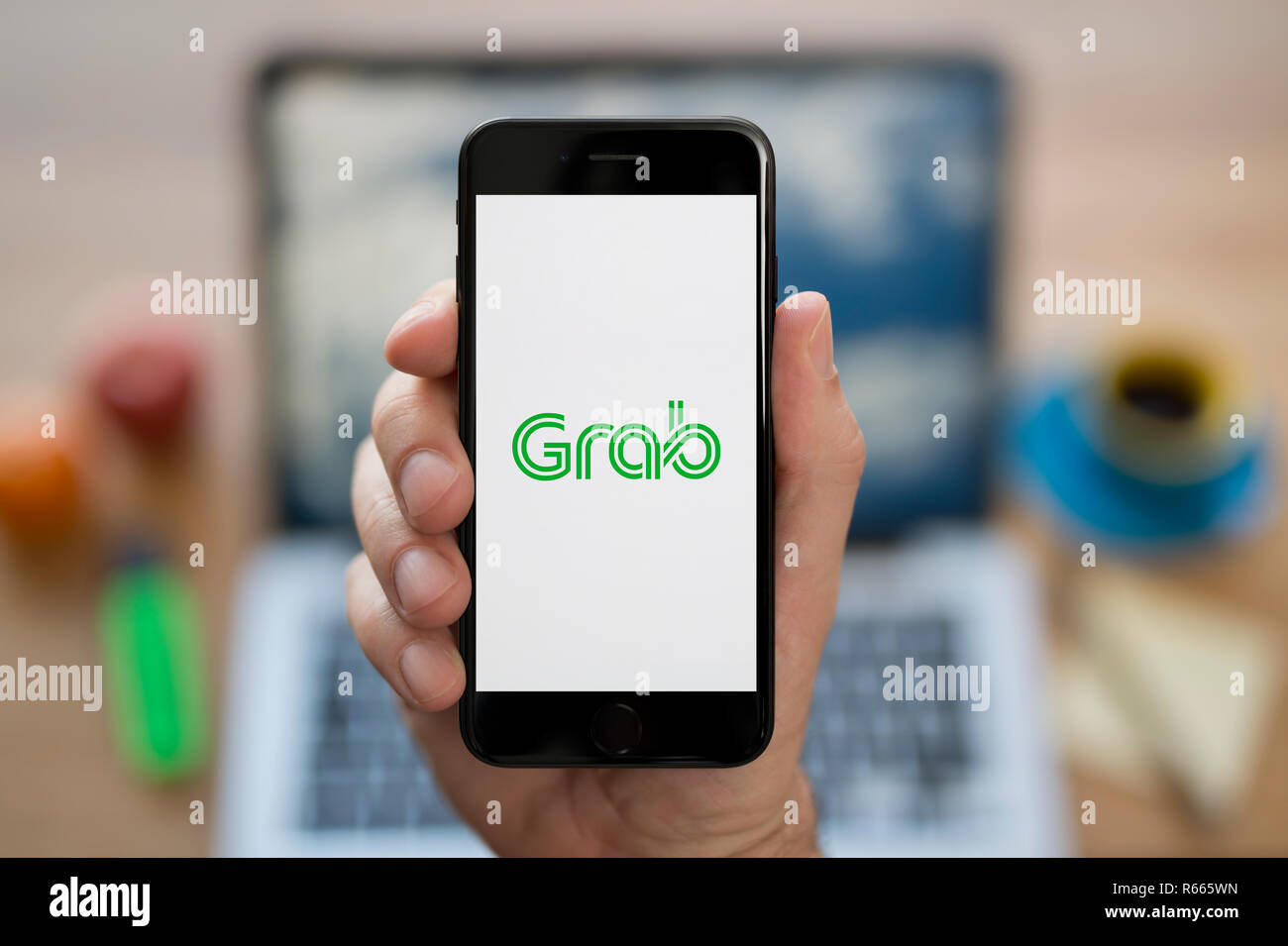A man looks at his iPhone which displays the Grab logo, while sat at his computer desk (Editorial use only). Stock Photo