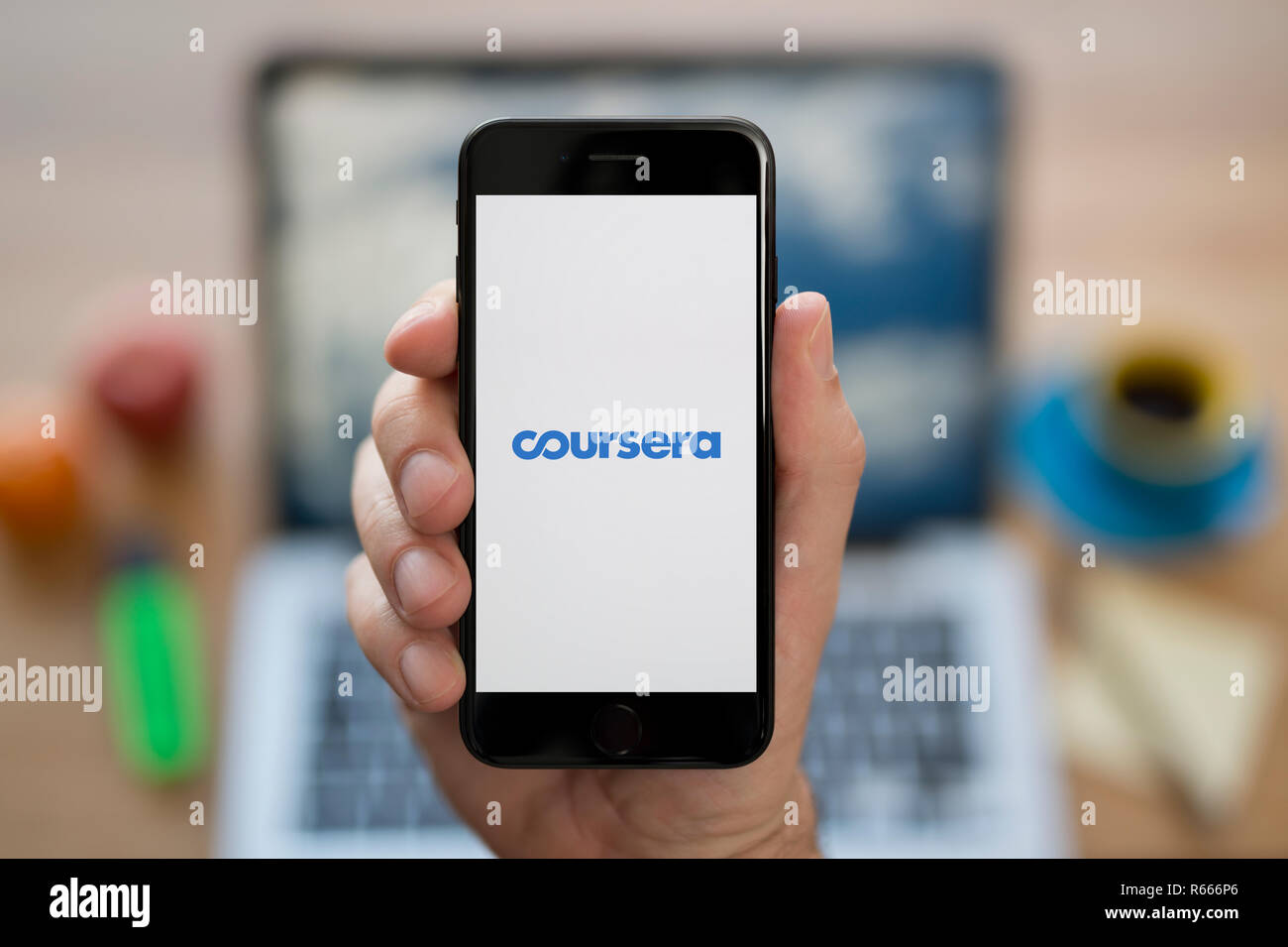 A man looks at his iPhone which displays the Coursera logo, while sat at his computer desk (Editorial use only). Stock Photo