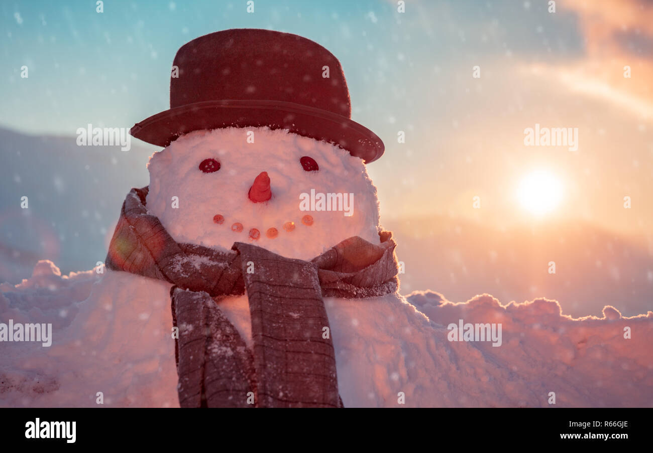 Photo of a cute snowman dressed in a stylish hat and scarf in a snowy frosty evening on a sunset background, happy winter holidays tradition Stock Photo