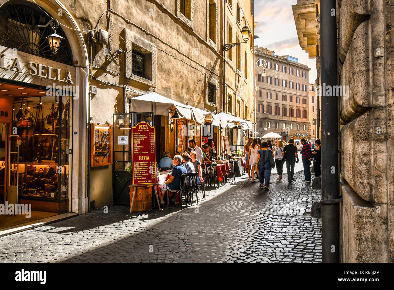 Tourists and local Italians enjoy afternoon shopping and dining at a sidewalk cafe patio on a narrow side street in the historic center of Rome Italy Stock Photo