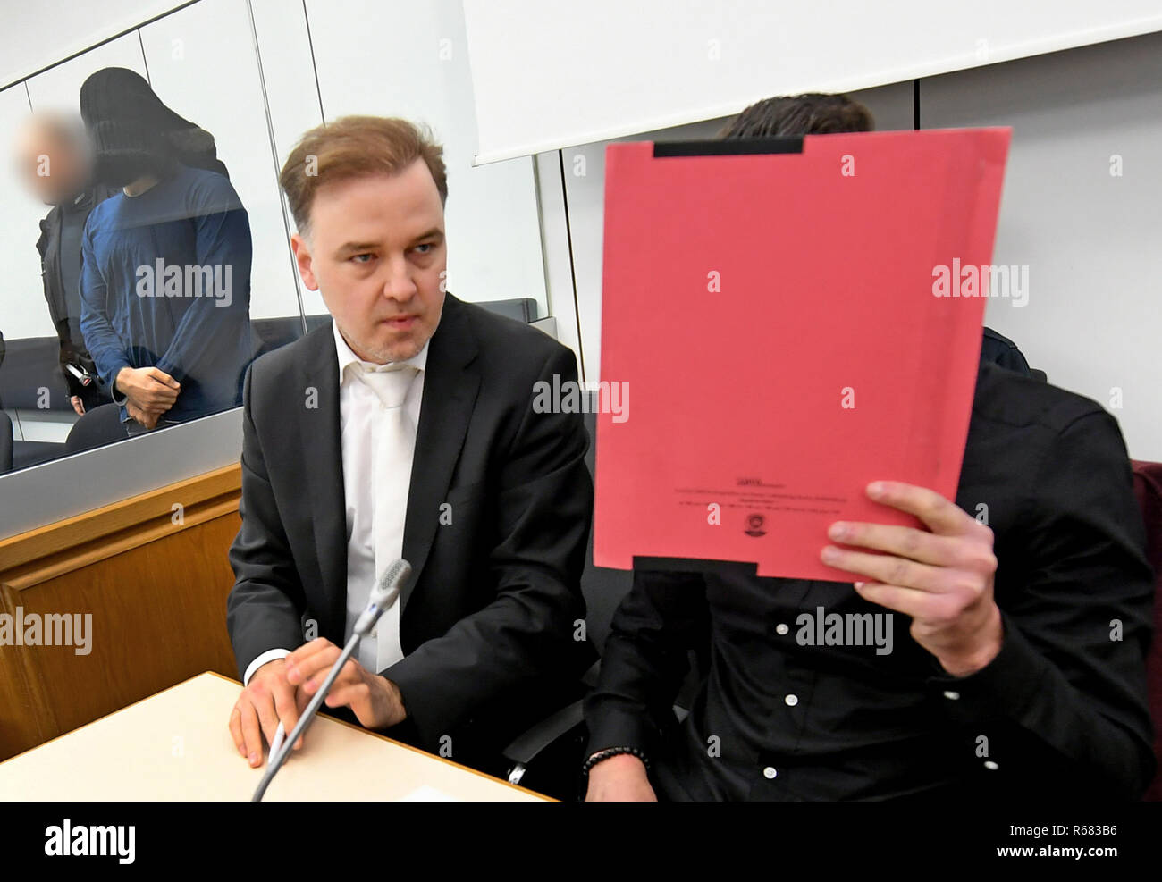 Celle, Lower Saxony. 04th Dec, 2018. A witness for the prosecution covers his face in a courtroom of the Higher Regional Court next to his defender Burkhard Benecken. The witness is to be heard from this Tuesday. He is one of the convicted juvenile assassins of the Sikh Temple in Essen in 2016 with three injured. In the background is Abu Walaa, alleged leader of the terrorist militia Islamic State (IS) in Germany. Walaa and four other alleged top Islamists on trial are said to have recruited volunteers for the IS. Credit: Holger Hollemann/dpa/Alamy Live News Stock Photo