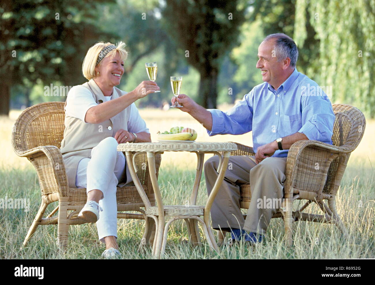Portrait, Outdoor, aelteres Paar sitzt auf Korbsesseln im Park und trinkt Sekt Stock Photo