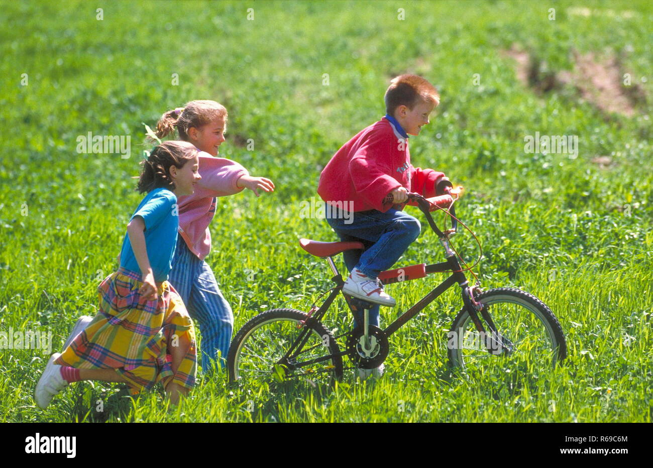 Parkszene, Gruppe von 3 Kindern im Alter von 6-10 Jahren, 2 Maedchen folgen zu Fuss einem Jungen auf dem Fahrrad durch eine Wiese Stock Photo