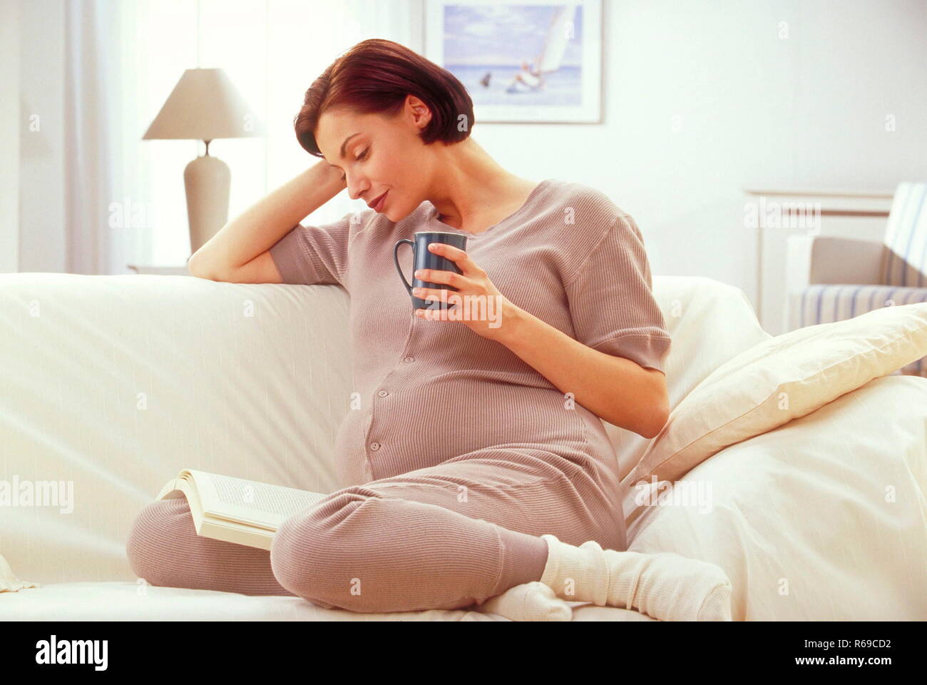 Portrait, hochschwangere Frau sitzt entspannt auf ihrem Sofa, liest ein Buch bei einer Tasse Tee Stock Photo