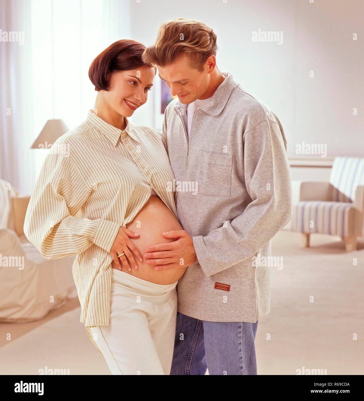 Portrait, Mann steht im Wohnzimmer und beruehrt den Bauch seiner hochschwangeren Frau Stock