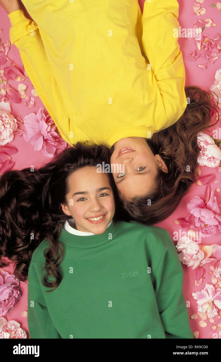 Portrait, 2 Maedchen mit langen braunen Haaren, 10 Jahre, liegen laechelnd flach auf einem mit rosa Blueten uebersaeten rosa Boden Stock Photo