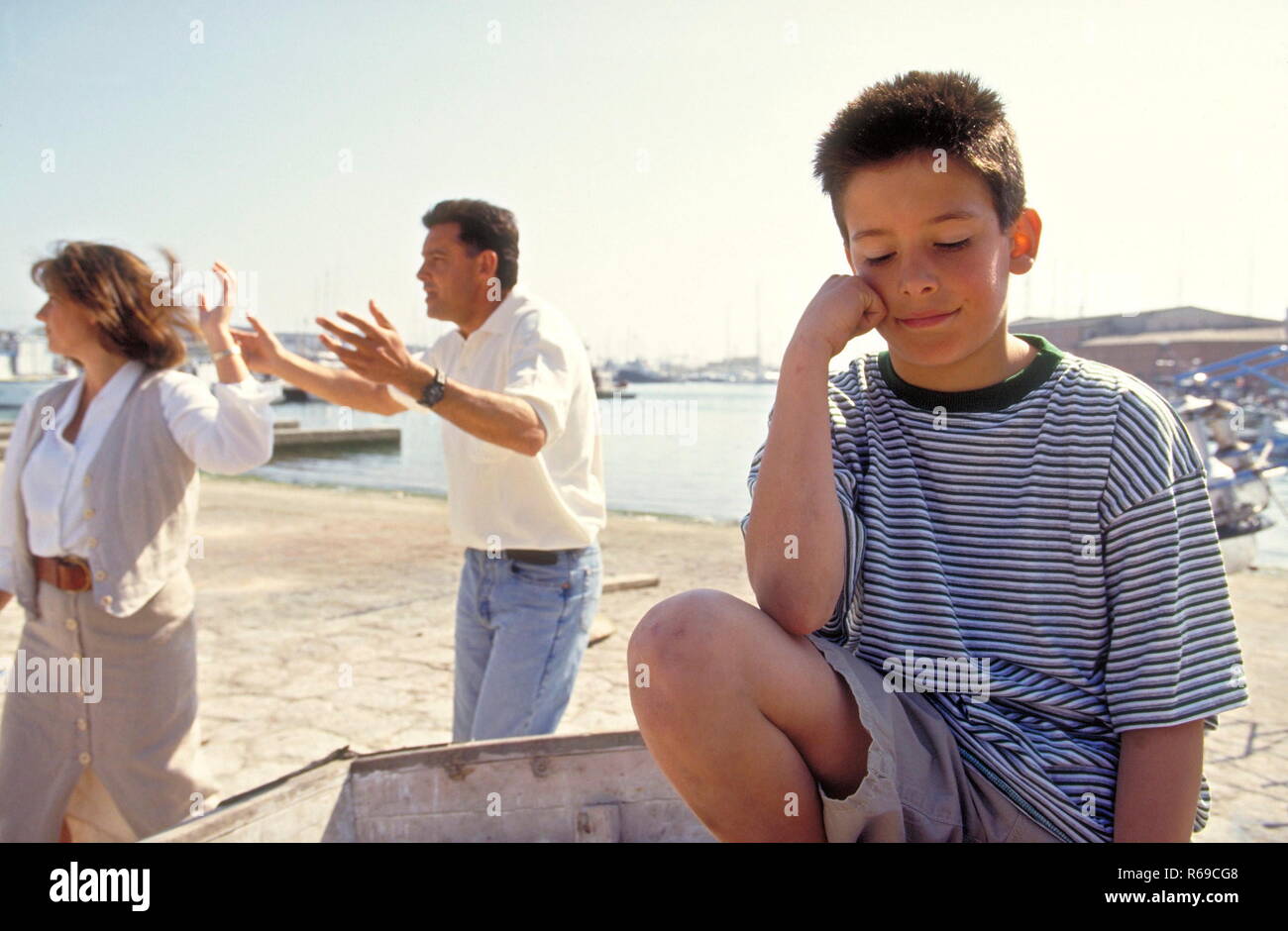 Portrait, Junge mit kurzen braunen Haaren sitzt traurig in einem Ruderboot, waehrend sich seine Eltern auf dem Kai lautstark streiten Stock Photo